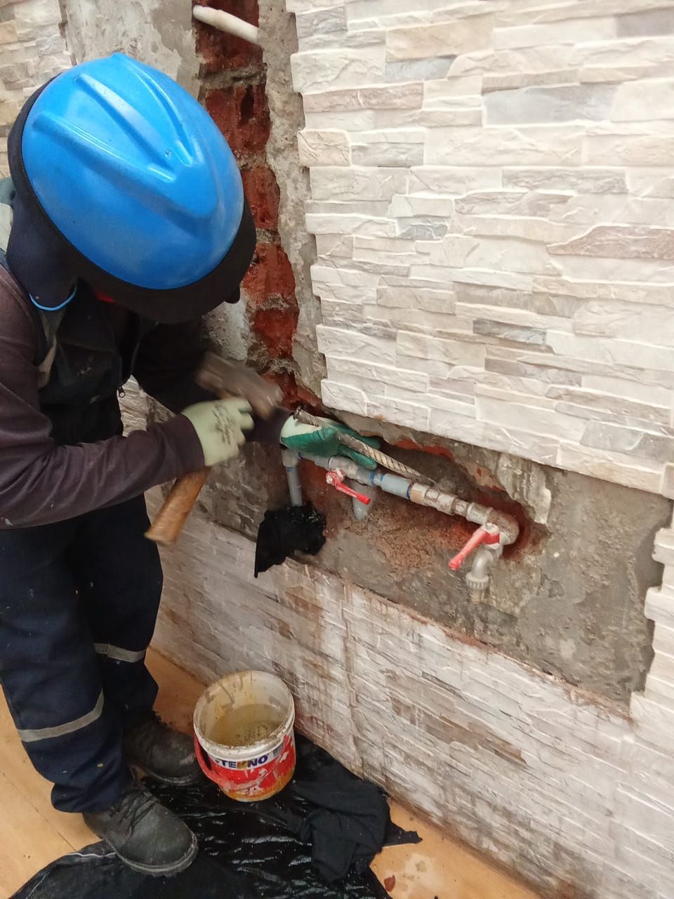 Persona con casco azul trabajando en una tubería en una pared de ladrillos. Hay un cubo y una lona negra.
