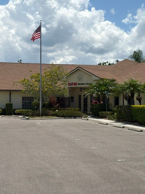 Single-story office building with American flag, red-tile roof, palm trees, and empty parking lot under cloudy sky