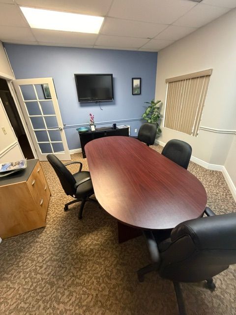 Conference room with oval wooden table, black chairs, wall-mounted TV, and window blinds.