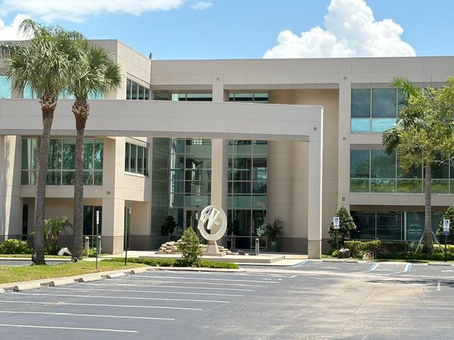 Modern office building entrance with palm trees, glass windows, and a sculpture in front