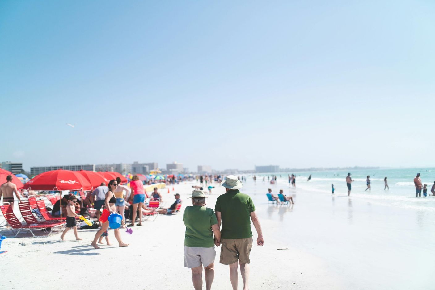 Crowded sunny beach with people under red umbrellas and two walkers in green shirts near the shoreline
