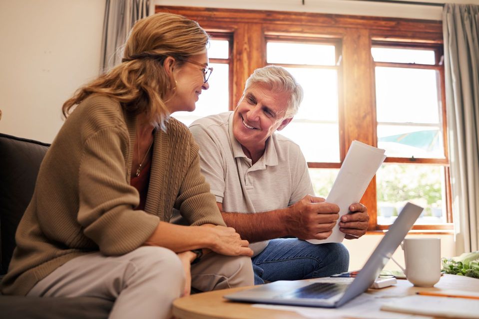 Two people talking on a couch, reviewing papers beside a laptop in a bright living room.