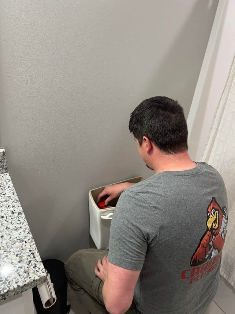 Man kneeling beside a toilet, reaching into the tank in a bathroom with gray walls.