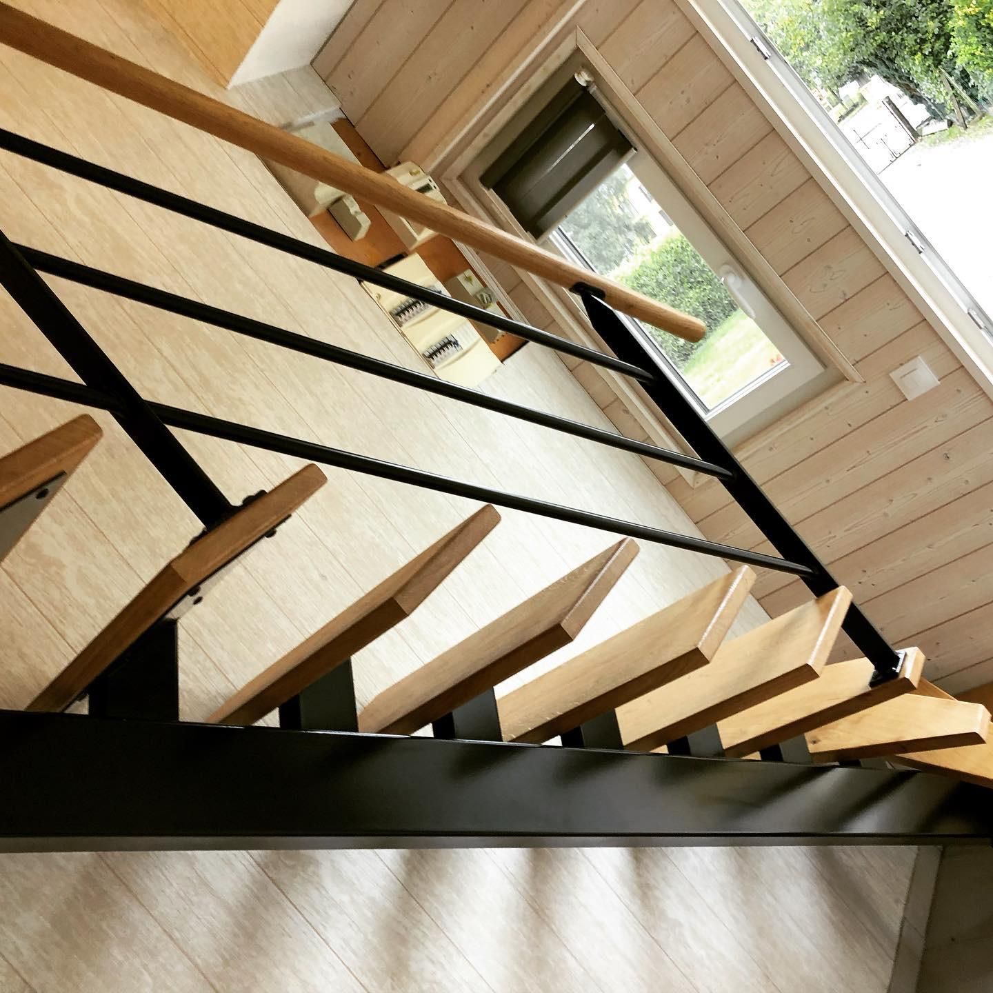Escalier en bois avec rampe en métal noir, menant vers le haut près d'une fenêtre.