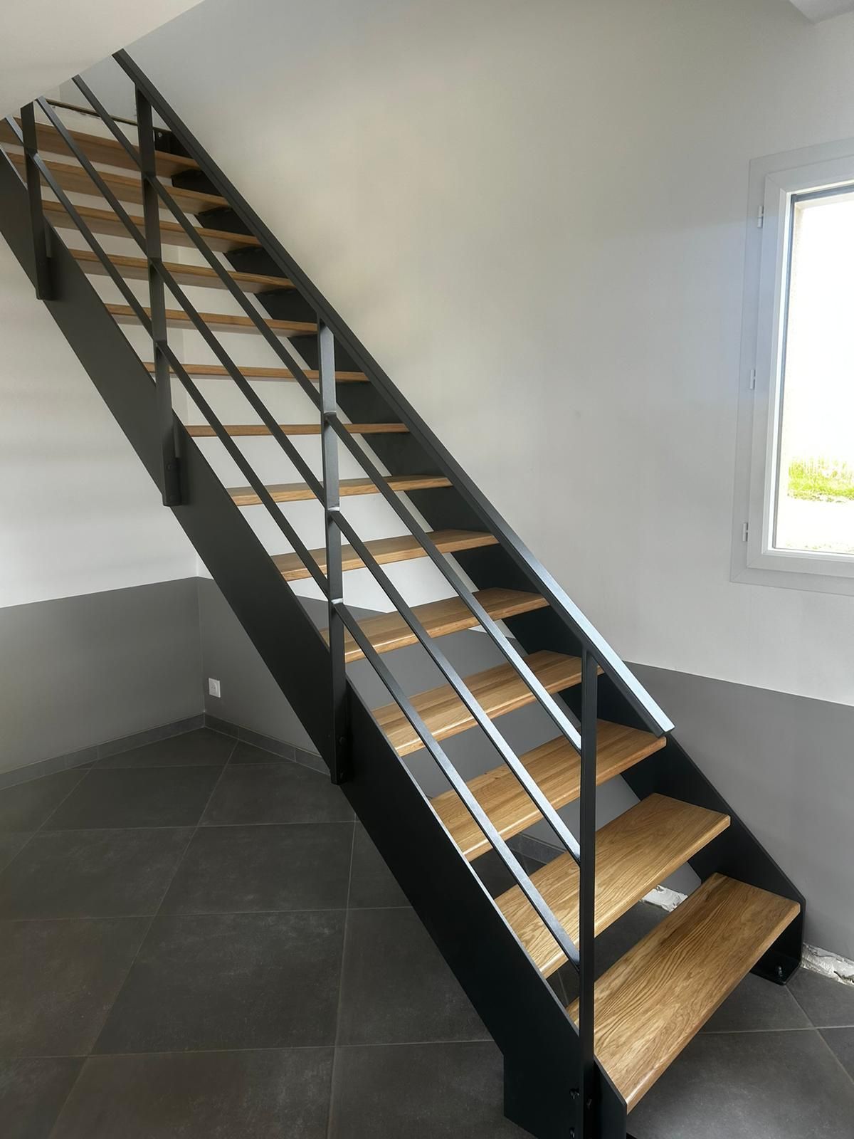 Escalier en métal avec marches en bois, rampe noire, dans une pièce avec sol en carrelage gris et murs blancs.