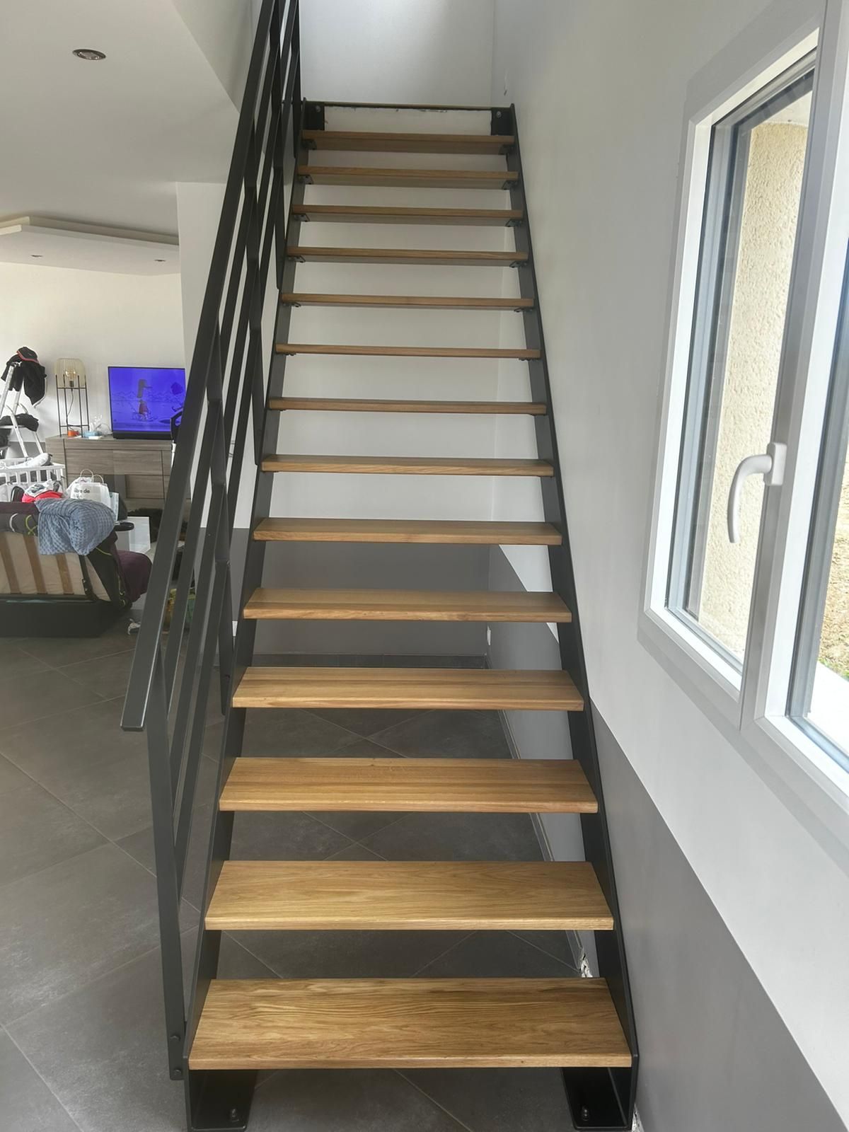 Escalier avec marches en bois et structure en métal noir sur fond de murs blancs et gris, à côté d'une fenêtre.