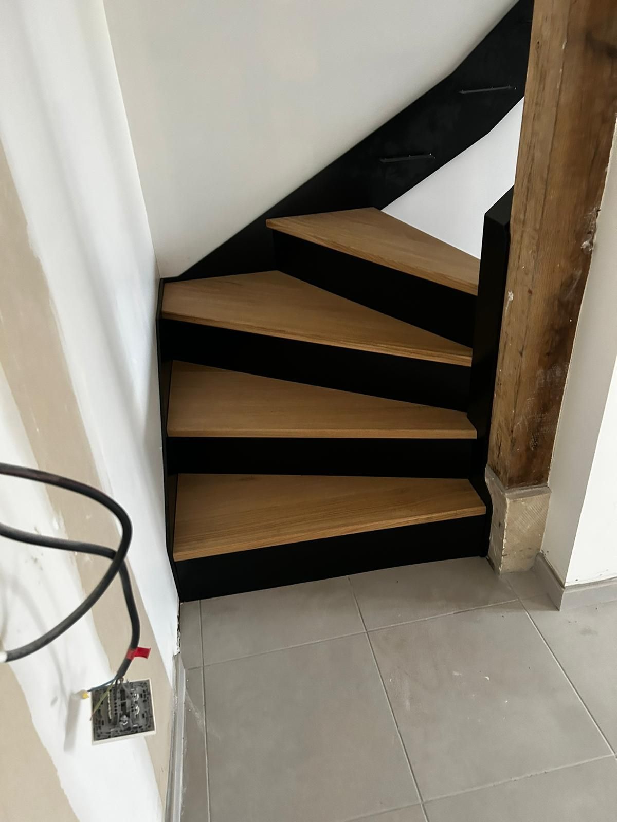 Escalier en bois avec garniture noire, montant dans un coin, à côté d'un pilier en bois.