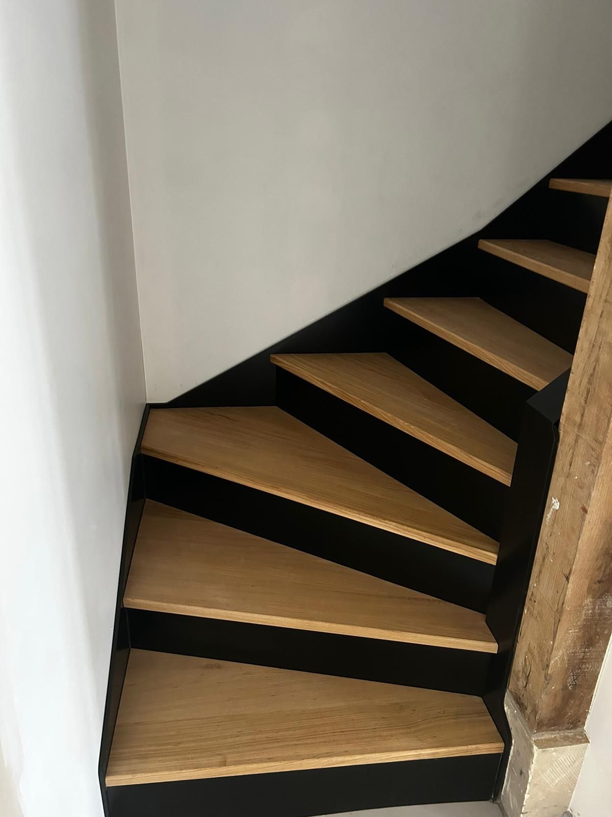 Escalier en bois avec marches claires et contremarches noires, menant vers le haut dans un coin contre des murs blancs.