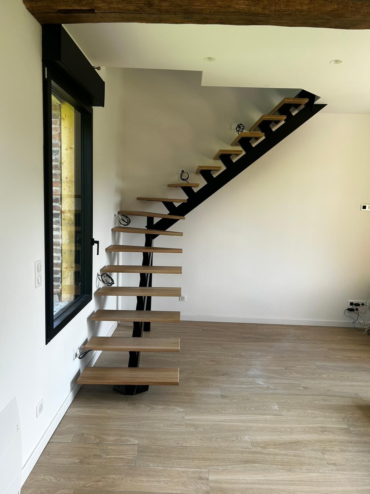 Escalier moderne en bois avec structure et supports en métal noir, à côté d'une fenêtre.