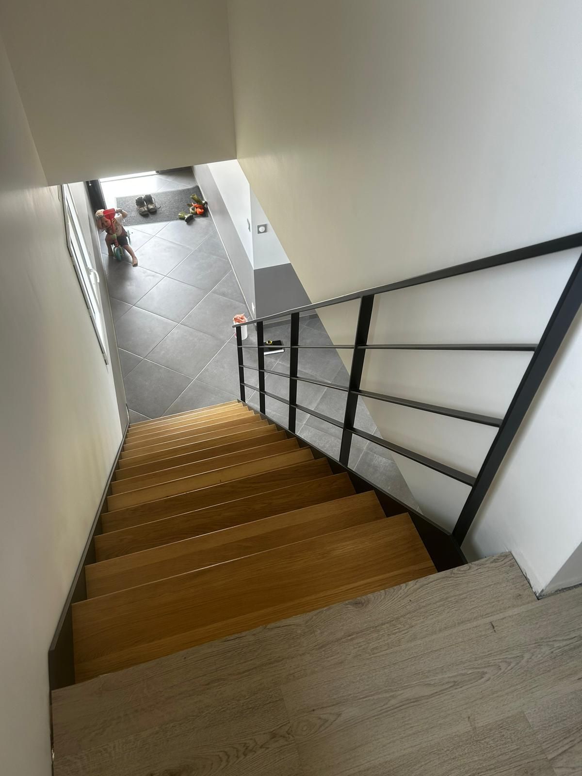 Escalier en bois avec rampe en métal noir et murs blancs, menant à un sol carrelé.