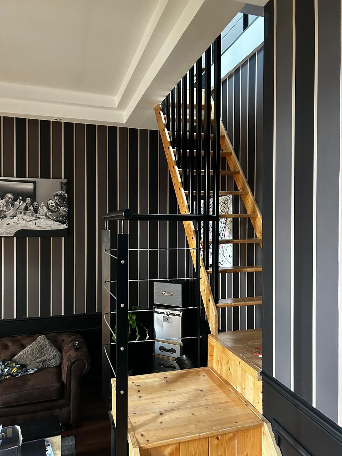 Escalier avec marches en bois et rampe noire sur fond de papier peint à rayures. Étagères métalliques noires en dessous.