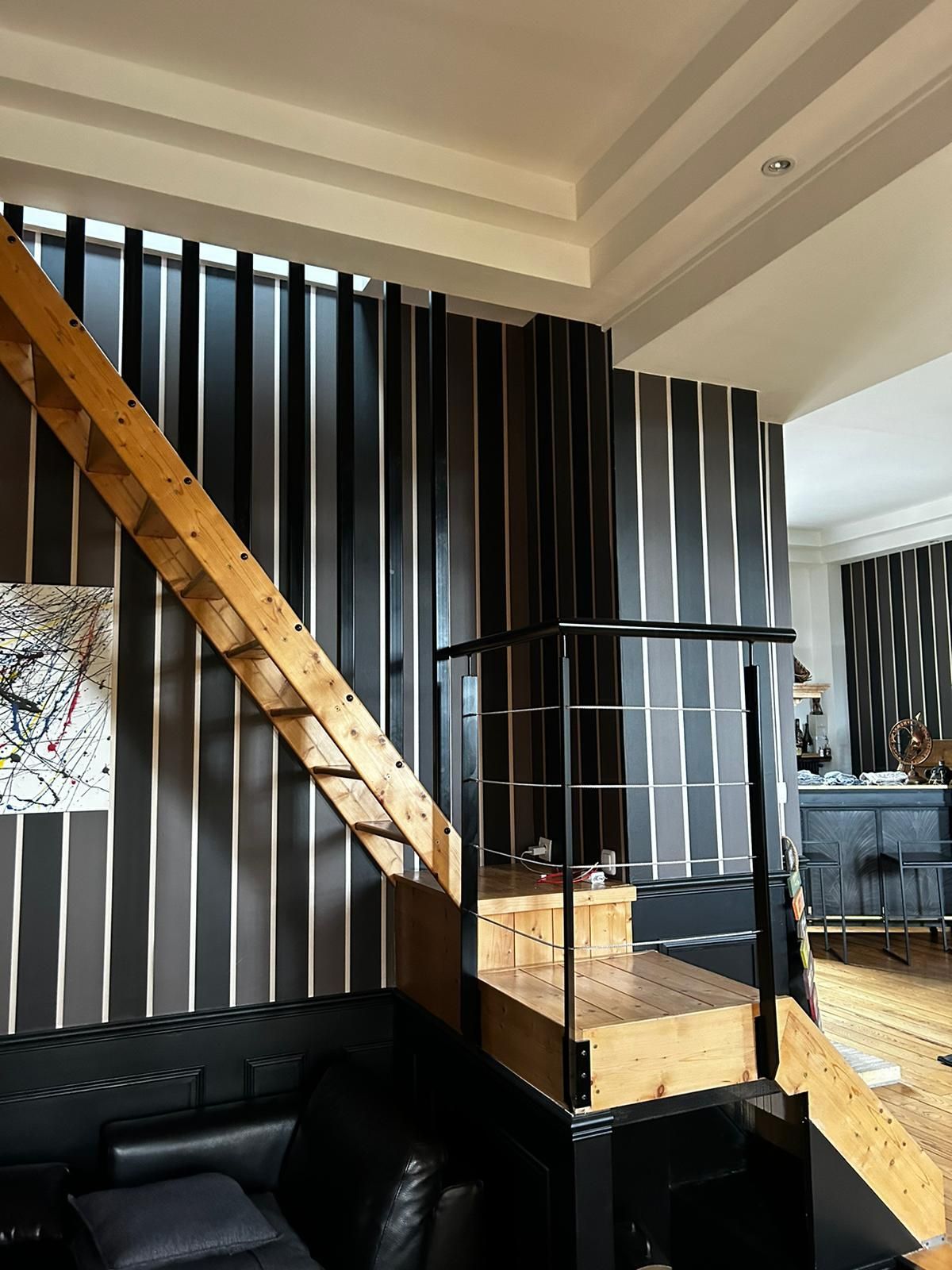 Escalier en bois avec rampe en métal noir, mur rayé noir et blanc et canapé noir.