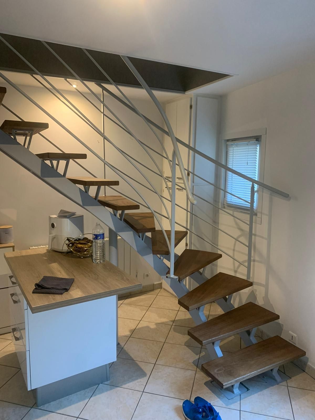 Escalier moderne en métal avec marches en bois. On aperçoit le comptoir de cuisine et une petite fenêtre.