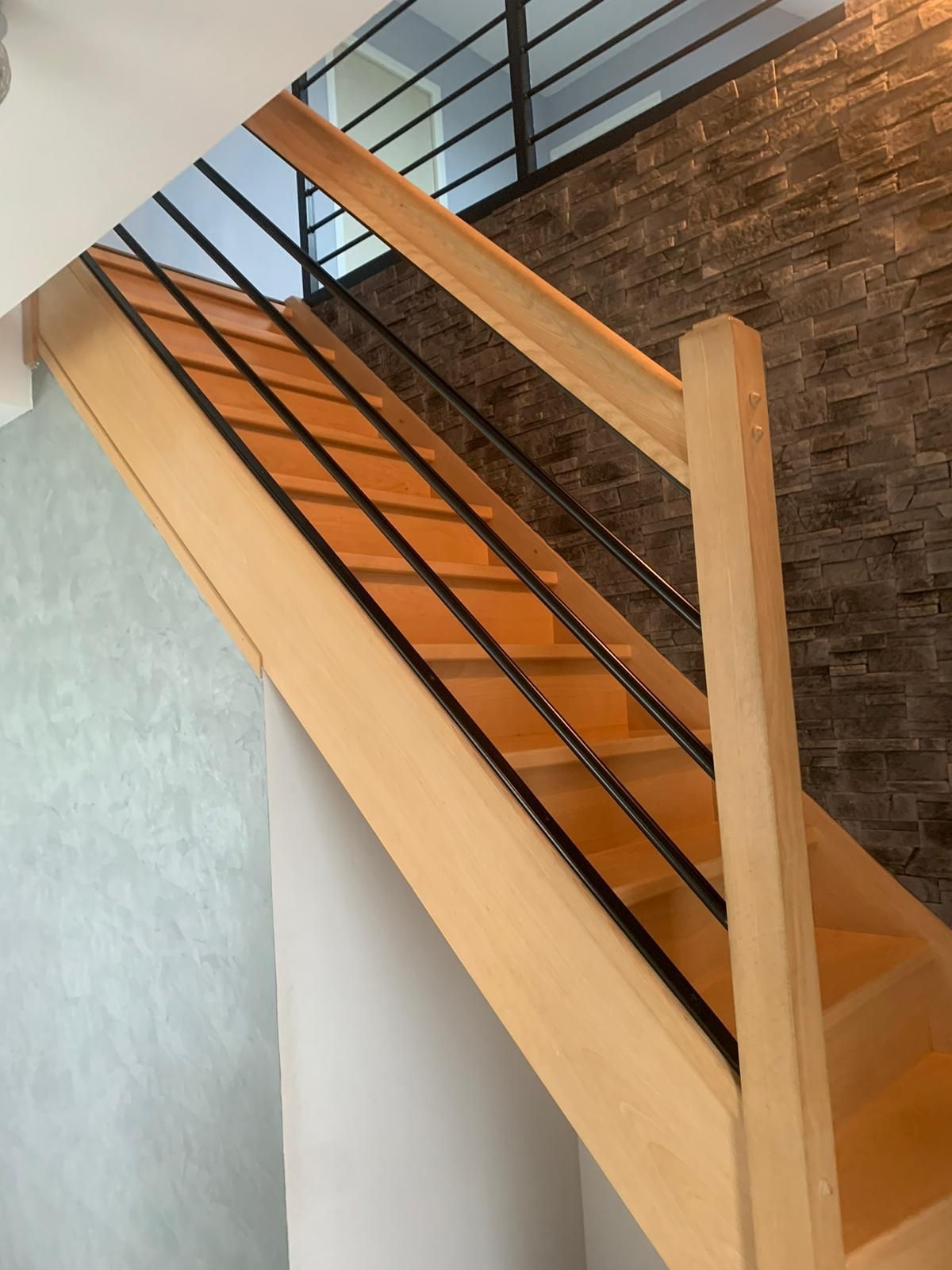 Escalier en bois avec rampes en métal noir, montant contre un mur de pierre texturé.