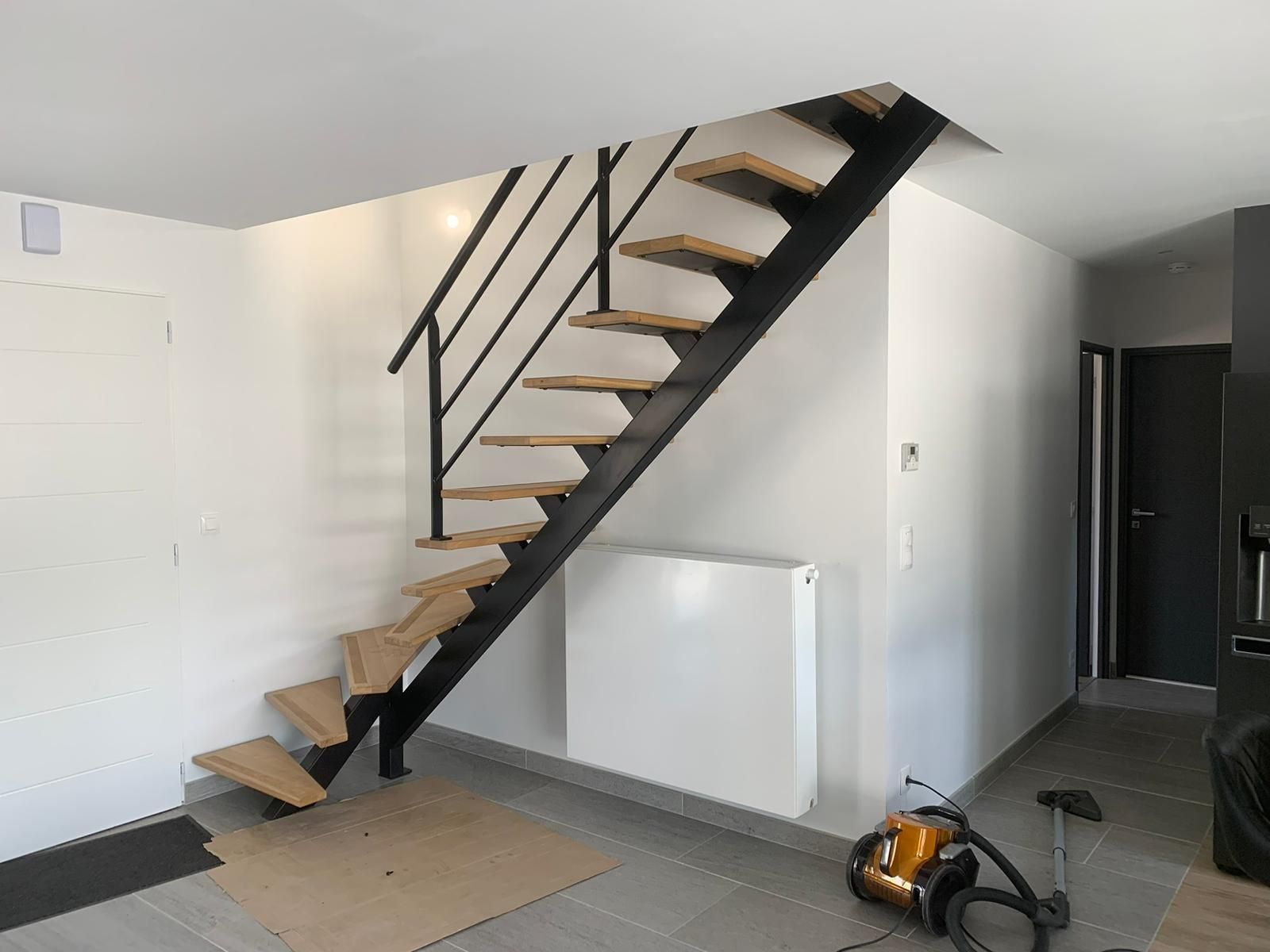 Escalier en métal noir avec marches en bois sur fond de murs blancs. Un aspirateur est posé au sol.