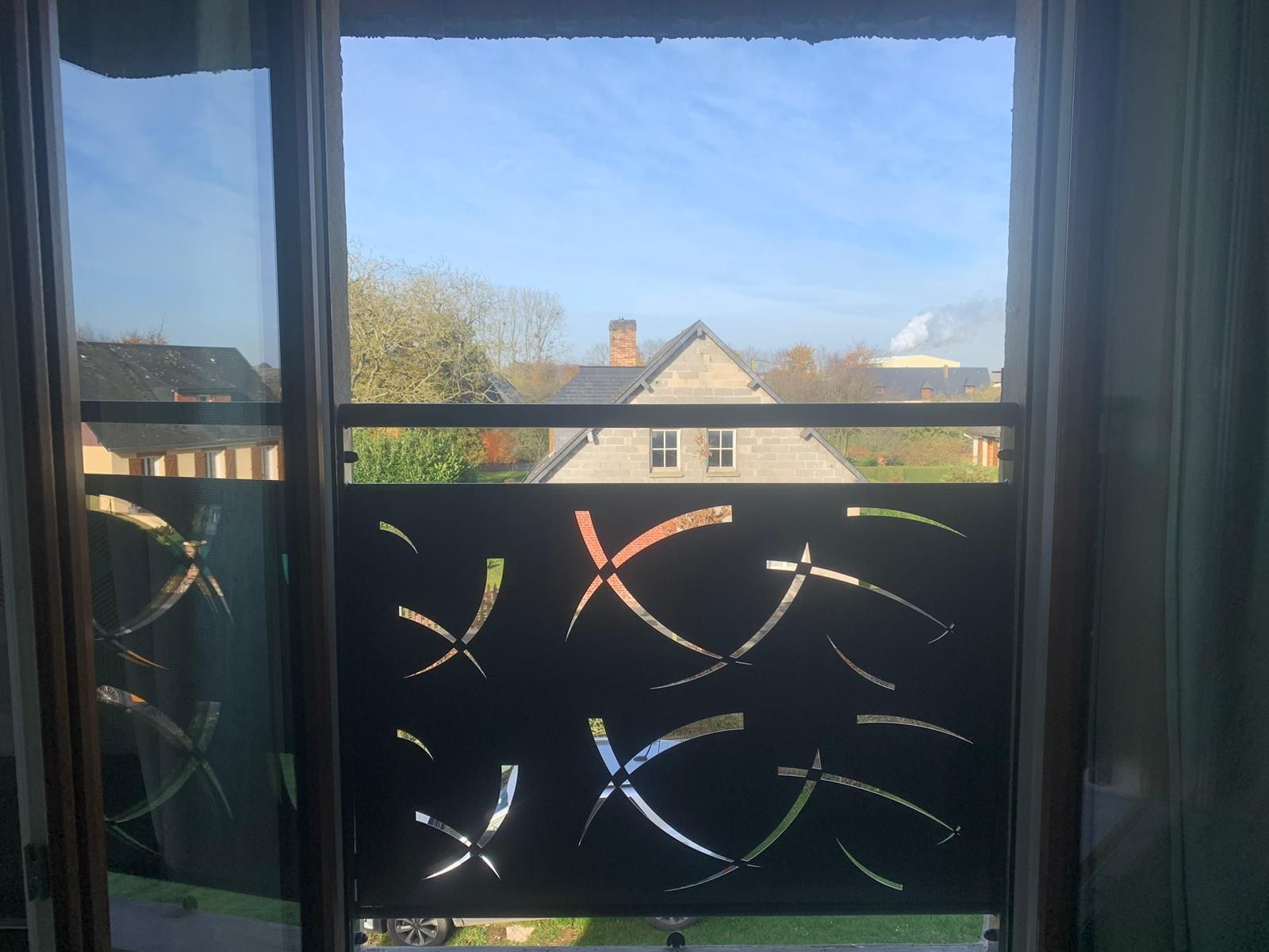 Vue d'une fenêtre avec un panneau décoratif noir orné de formes abstraites découpées. Maisons et ciel visibles en arrière-plan.