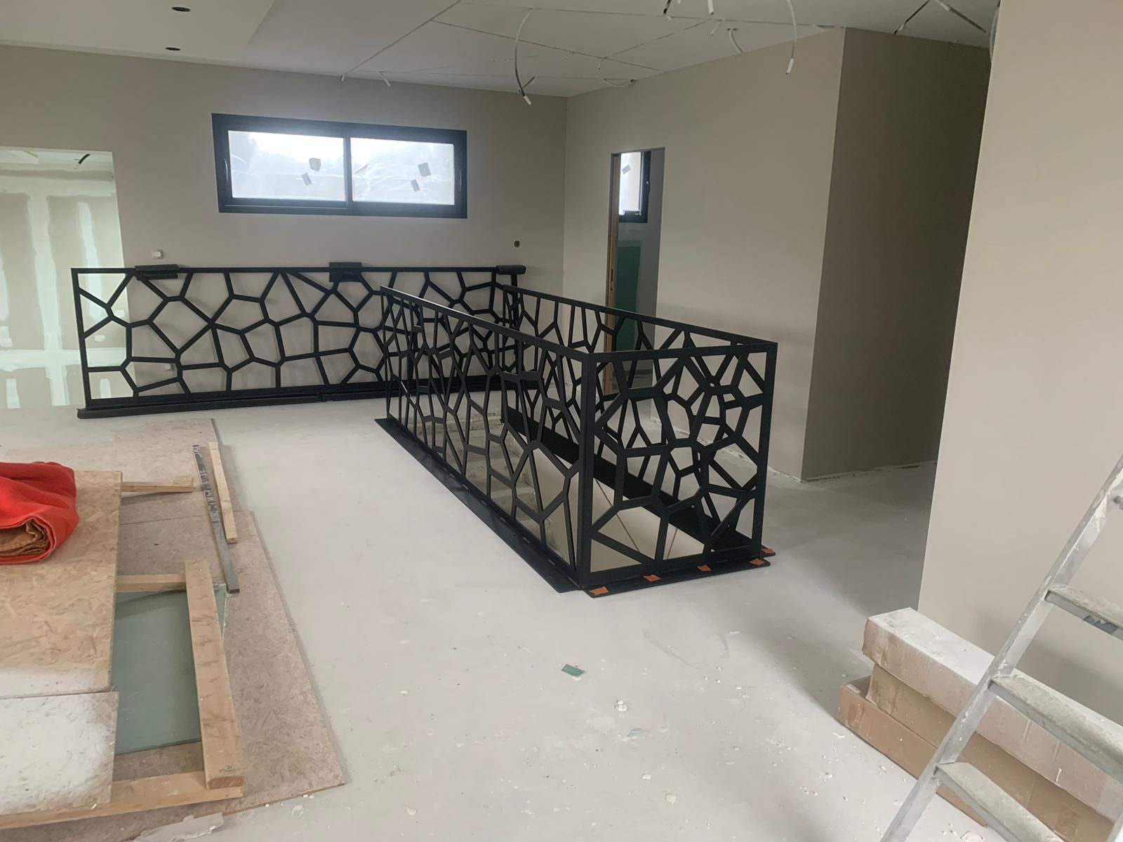 Garde-corps géométrique noir installé dans un intérieur lumineux, à côté d'une fenêtre et d'une entrée. Travaux en cours.