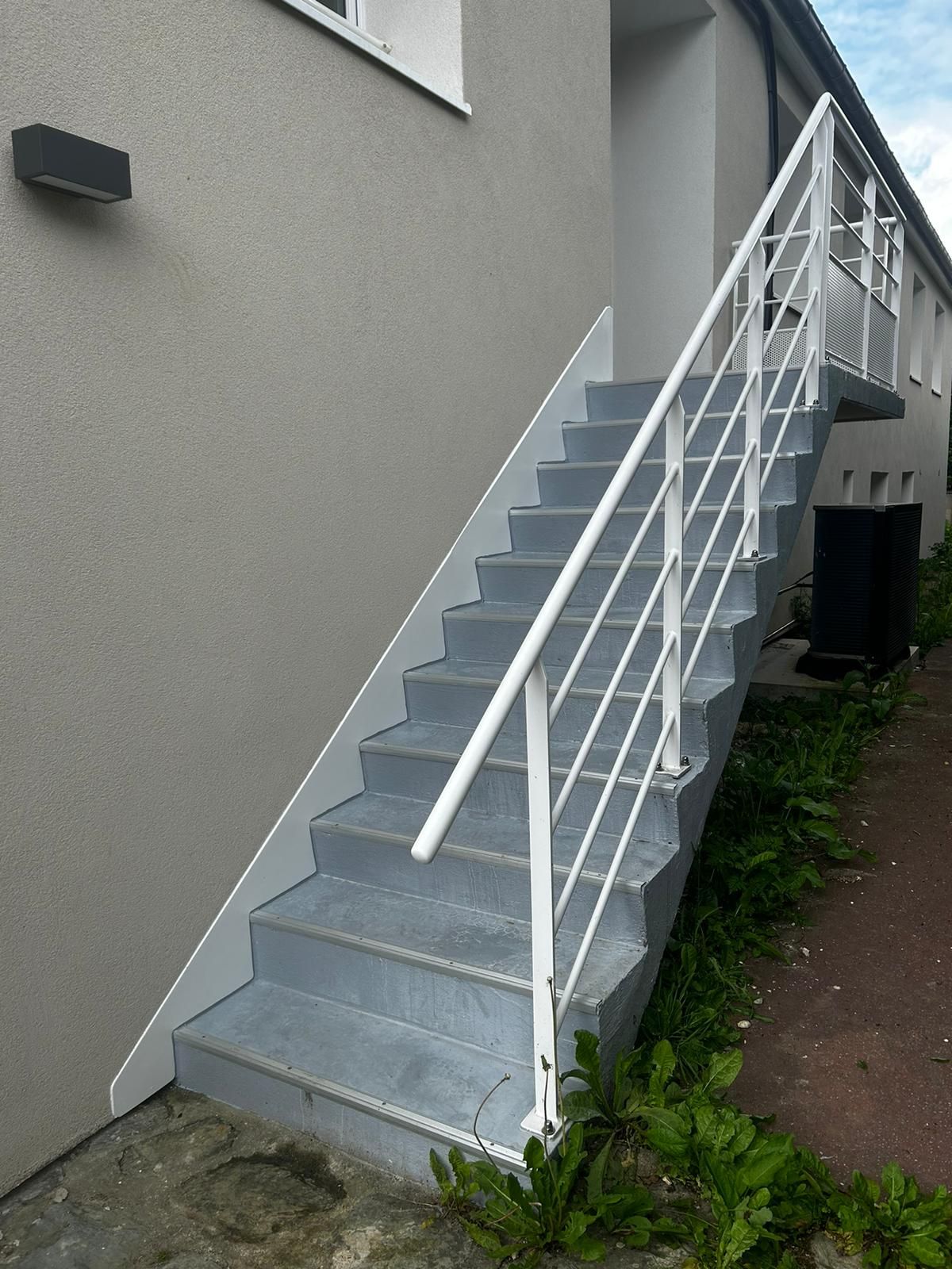 Escalier extérieur en métal peint en gris avec rampe blanche sur fond de bâtiment gris clair.