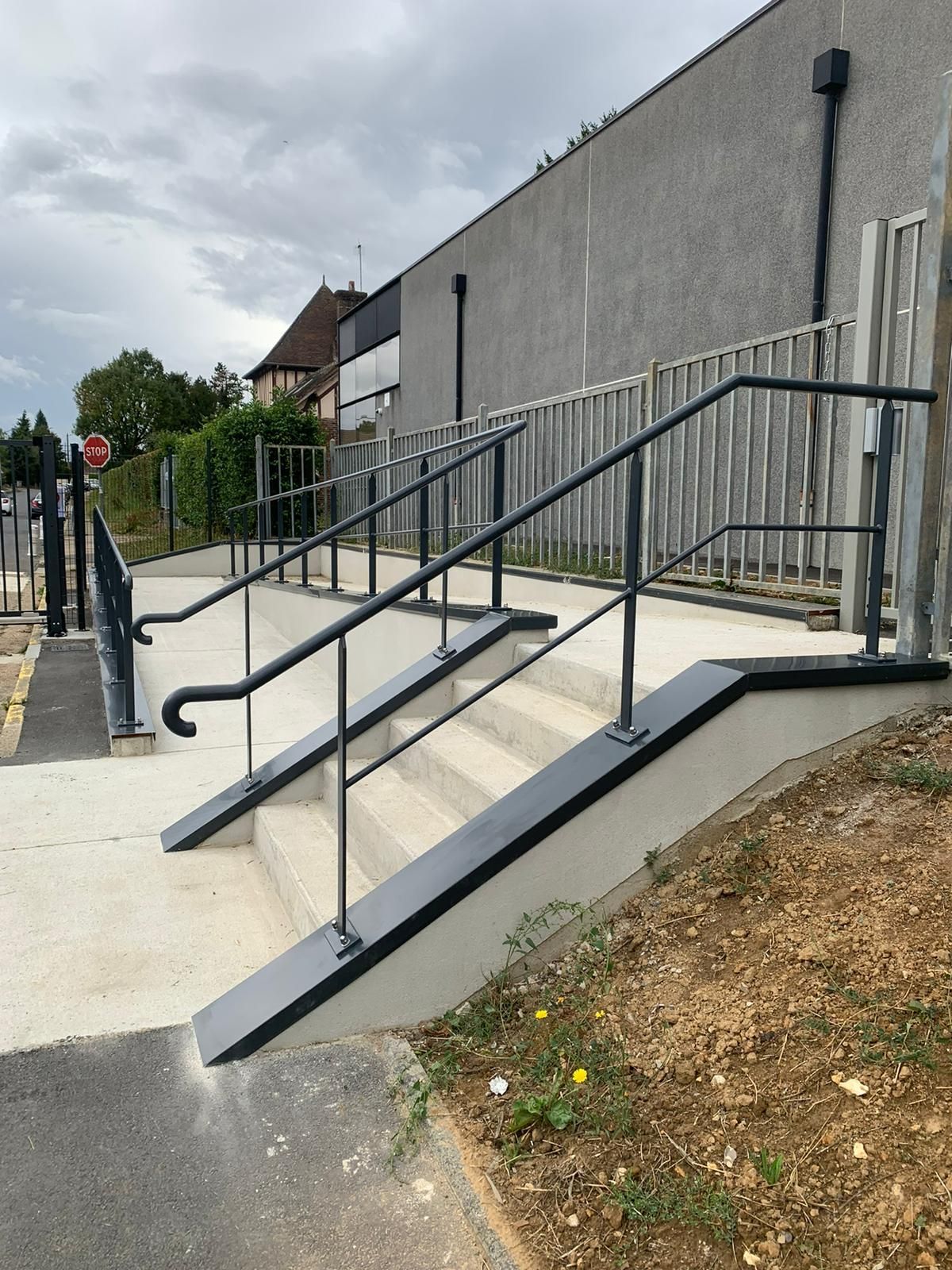 Escalier avec rampe et main courante noires, permettant un accès accessible à côté d'un bâtiment et d'une clôture.