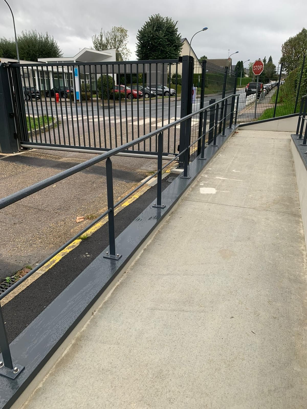 Rampe avec main courante près d'une clôture et d'un portail. Un chemin pavé mène à la rue. Temps couvert.