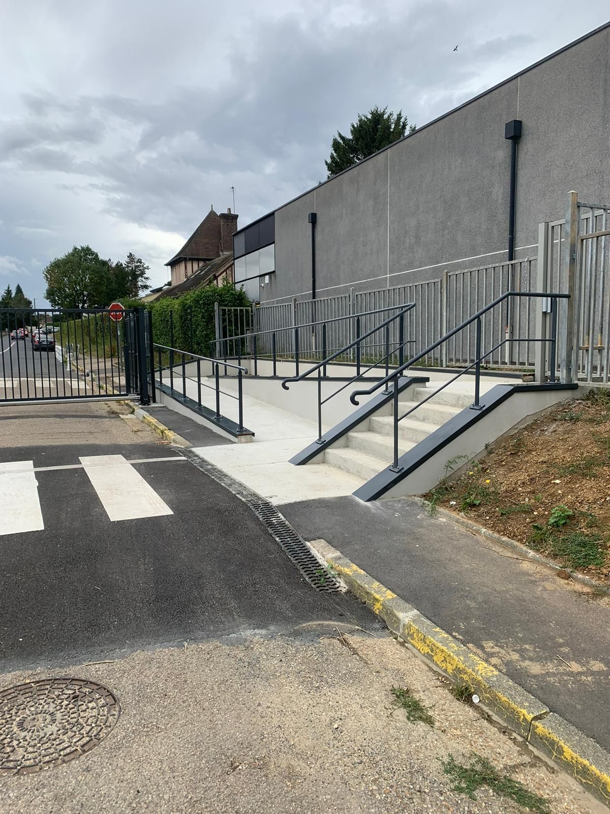 Rampe et escalier d'accès à un bâtiment à côté d'un passage piéton. Mur gris, garde-corps noirs, ciel couvert.