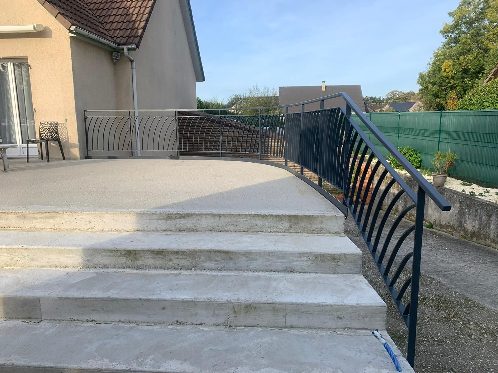 Marches en béton avec rampe en métal noir menant à un patio avec clôture beige ; maison en arrière-plan.