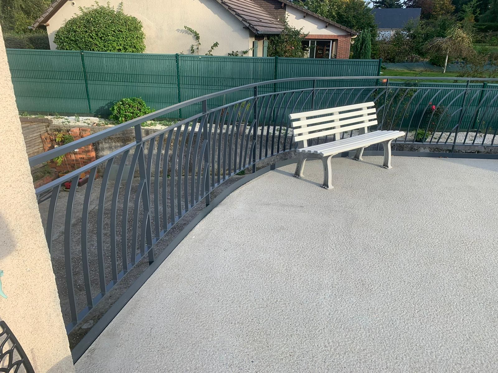 Un patio en béton avec une balustrade en métal courbée et un banc, derrière une clôture verte.