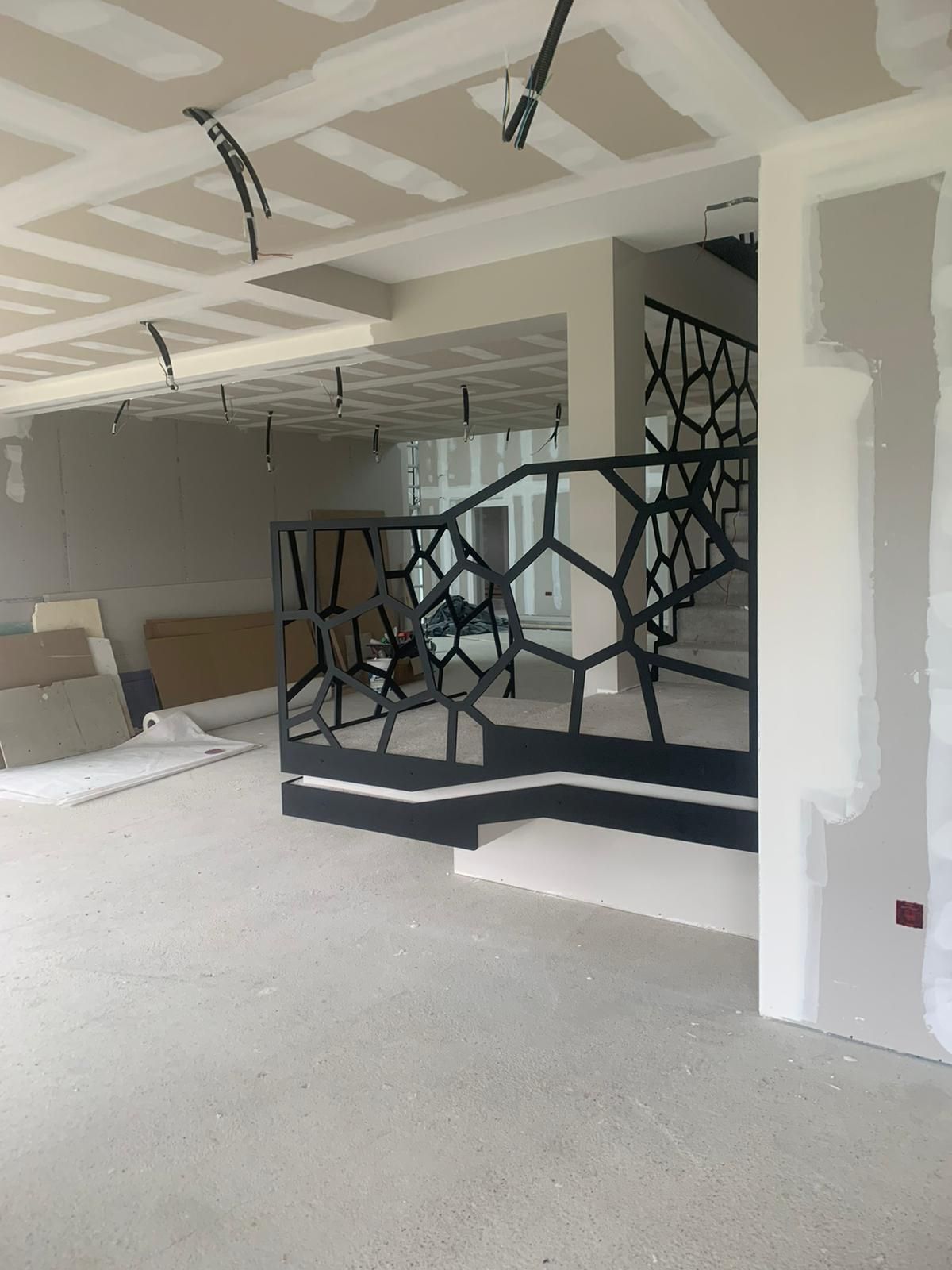 Vue intérieure de la construction : garde-corps géométrique noir, murs non finis, cloisons sèches, plafond et fils exposés.