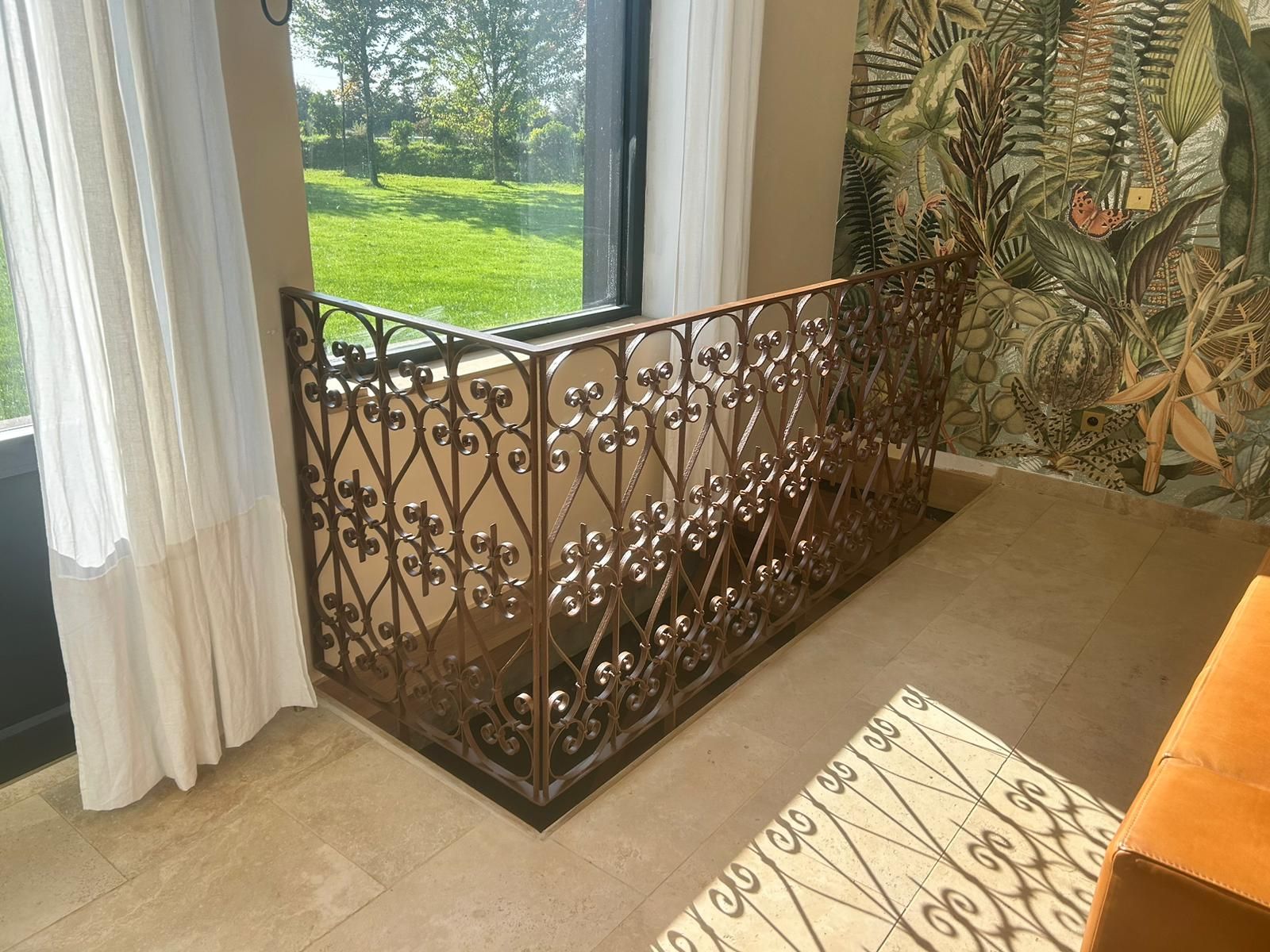 Rampe d'escalier en fer forgé à motif floral, à côté d'une fenêtre donnant sur une pelouse verte.