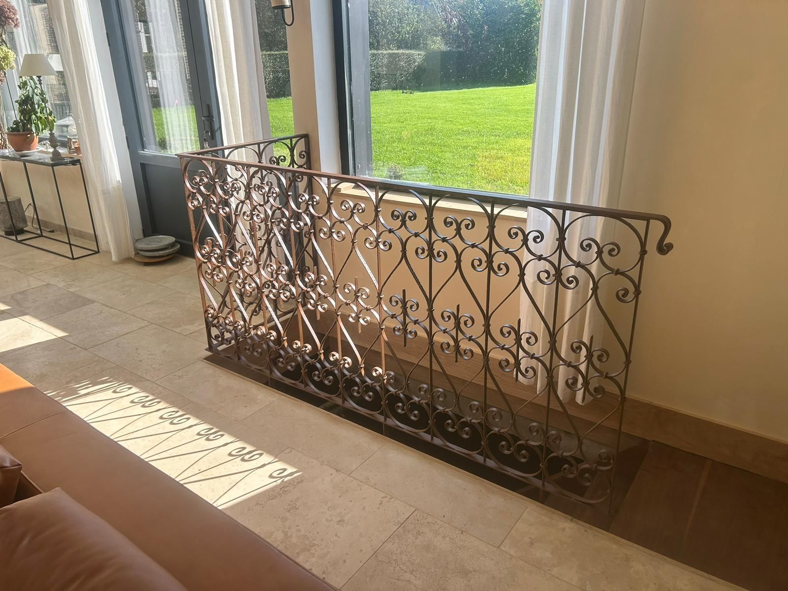 Rampe en fer forgé à volutes sur sol carrelé beige, à côté d'une fenêtre avec vue sur une pelouse.