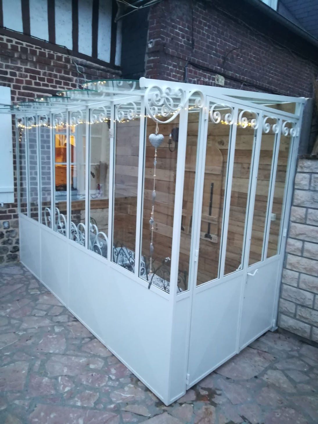 Véranda blanche aux parois vitrées, attenante à une maison en briques. Intérieur visible, patio en pierre.