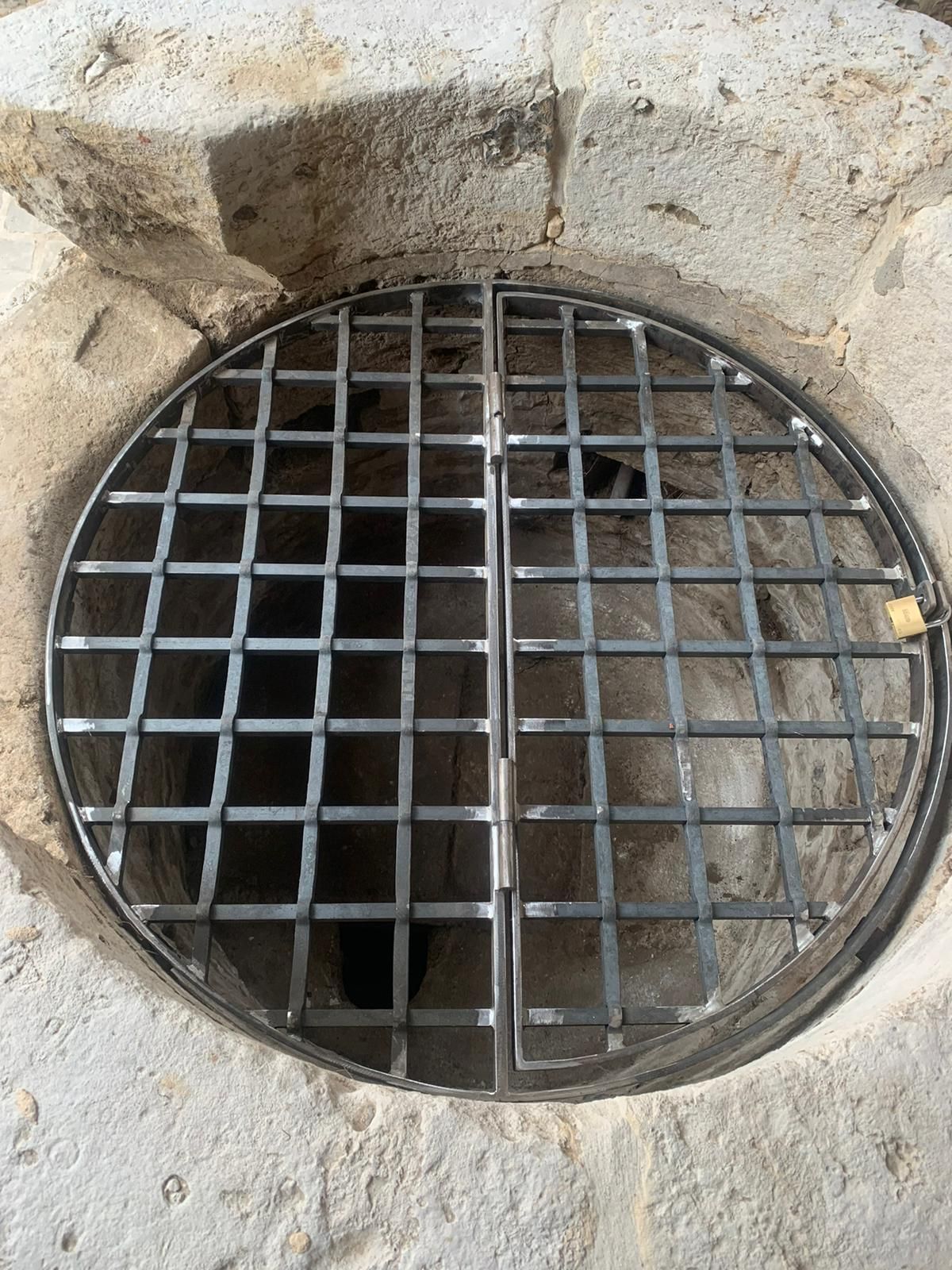Une grille métallique ronde recouvre l'ouverture d'un puits sombre encastré dans la pierre.