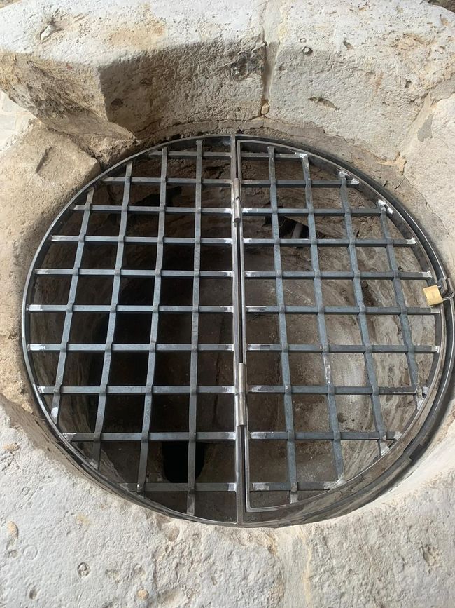 Une grille métallique ronde recouvre l'ouverture d'un puits sombre encastré dans la pierre.