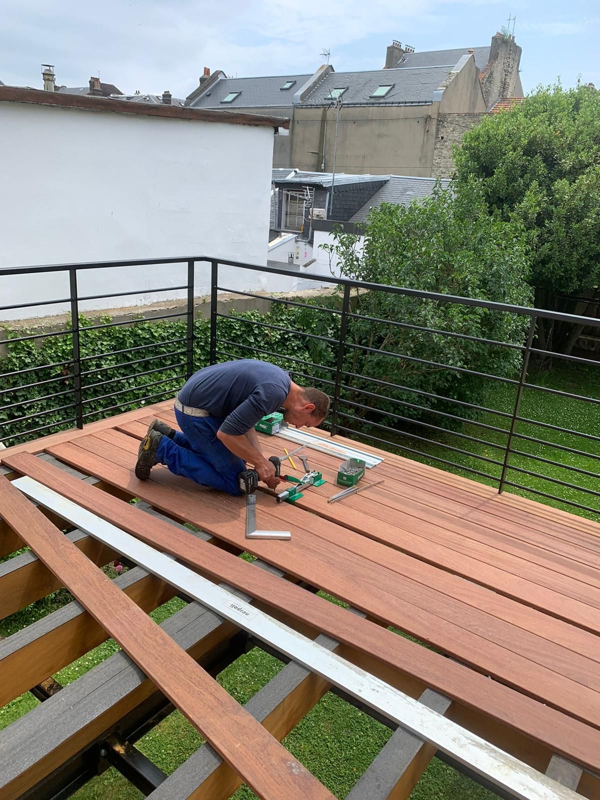 Un homme s'agenouille et utilise une perceuse pour installer une terrasse en bois sur un patio extérieur avec une balustrade noire.