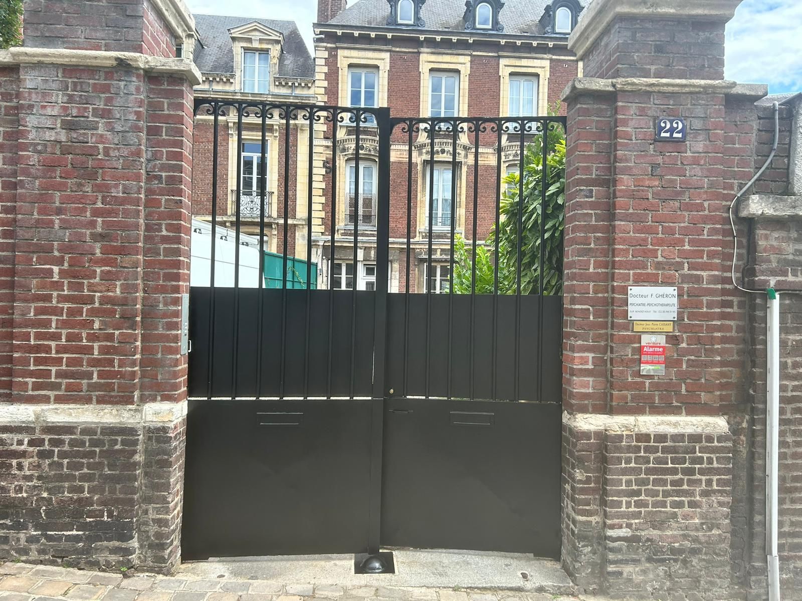 Portail noir entre des piliers en briques, menant à un bâtiment avec fenêtres. Le bâtiment présente un style architectural européen.