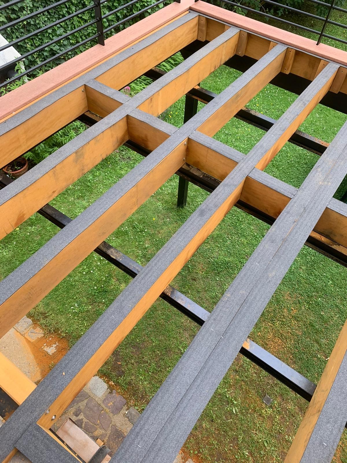 Structure de terrasse avec poutres en bois, supports noirs et platelage composite, posée sur une zone herbeuse.