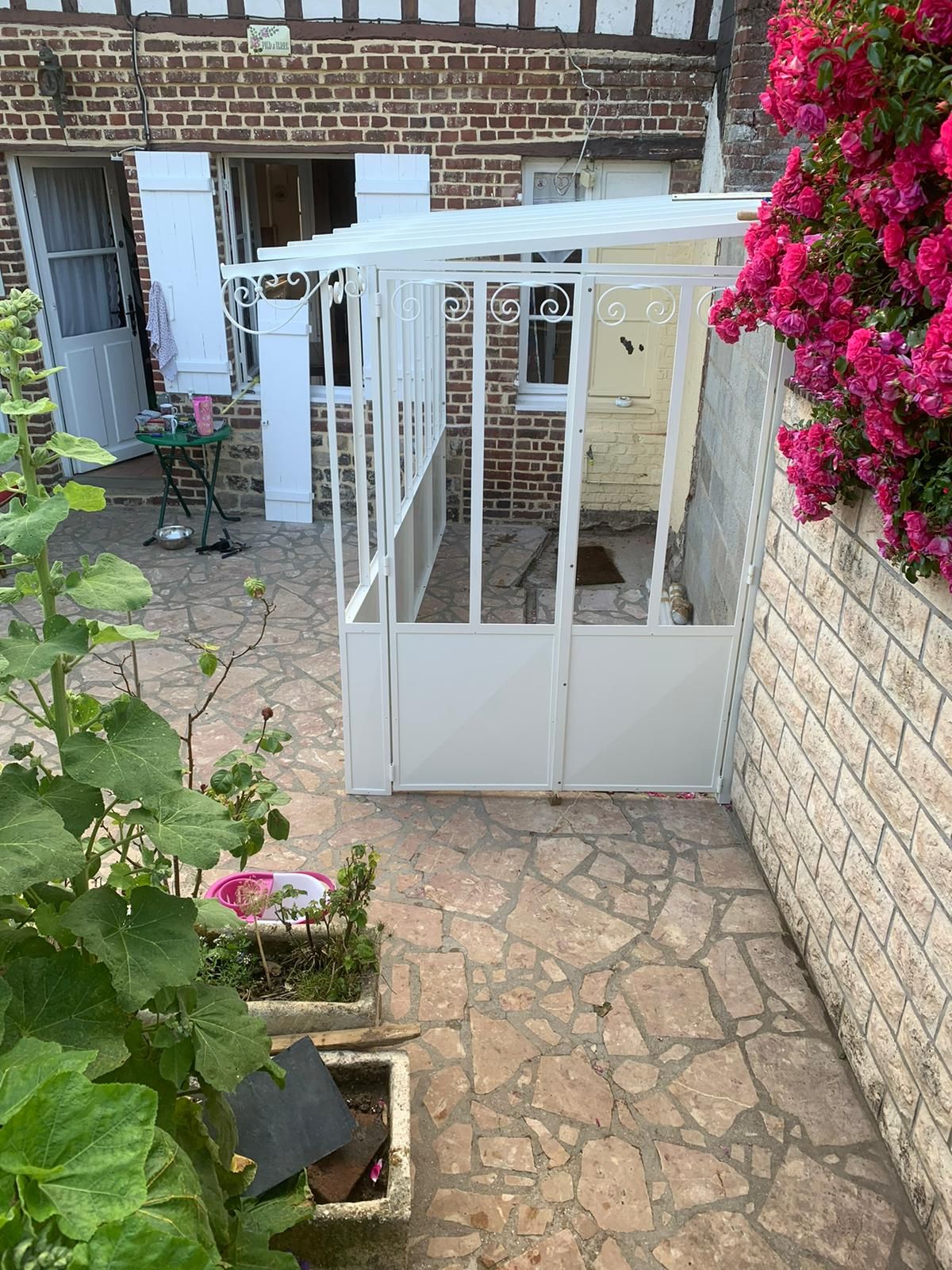 Espace patio blanc clos à côté d'un bâtiment en brique et pierre, avec des vignes roses fleuries.