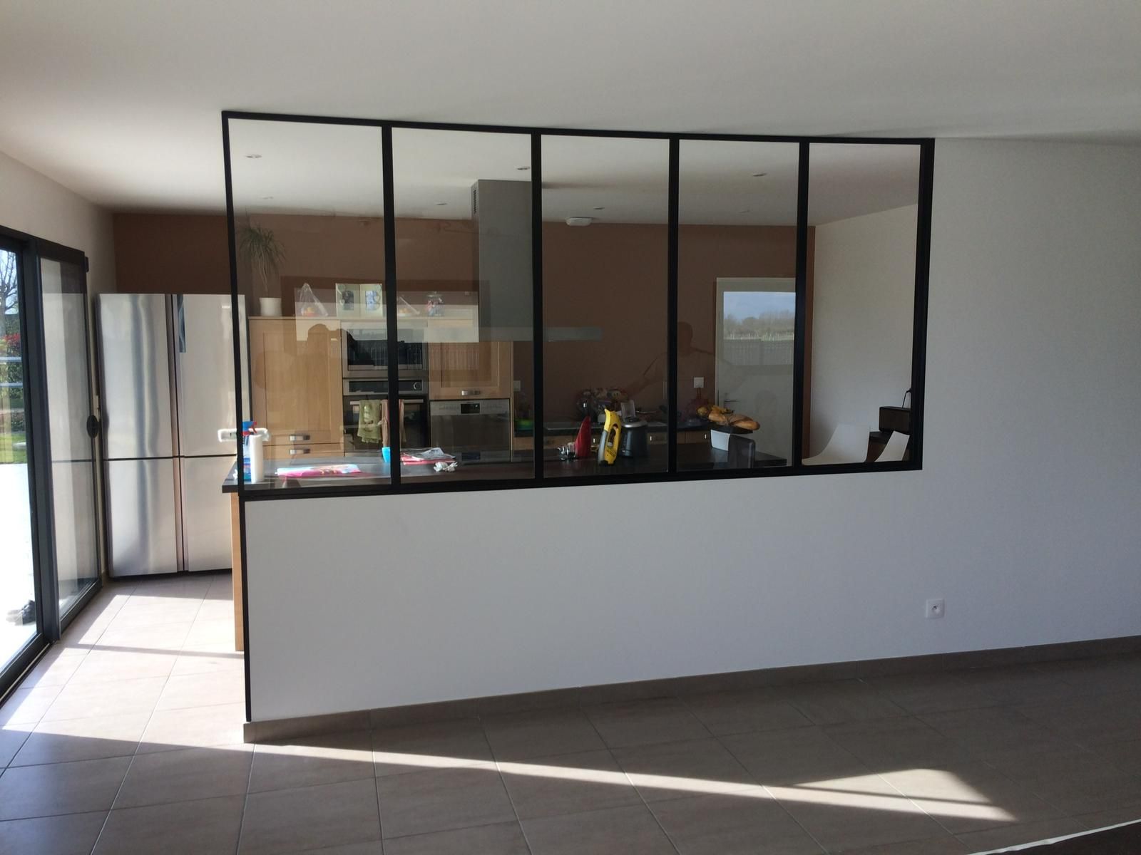 Intérieur avec mur blanc, fenêtres à cadre noir donnant sur la cuisine, réfrigérateur visible, sol carrelé.