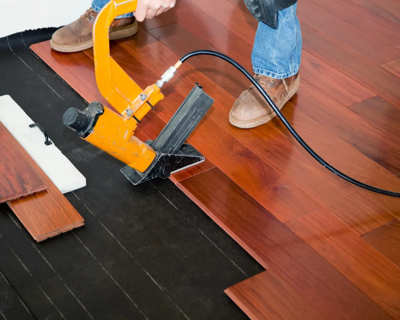 Person installing a reddish-brown wood floor with a yellow floor stapler on black underlayment