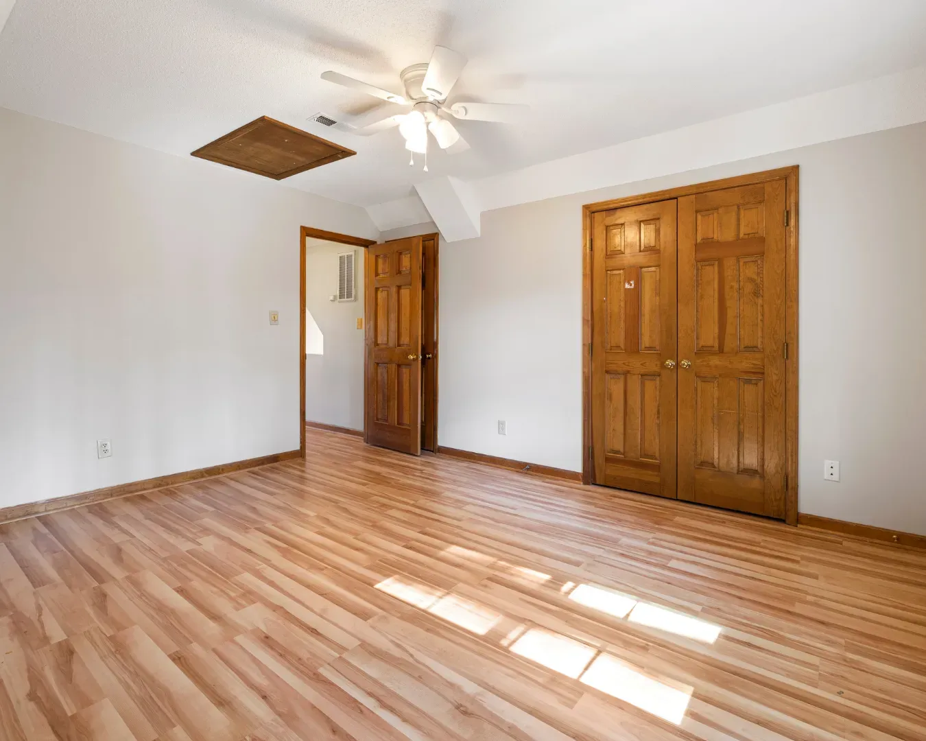 Empty bedroom with hardwood floors, white walls, ceiling fan, and two wooden closet doors open to a hallway.