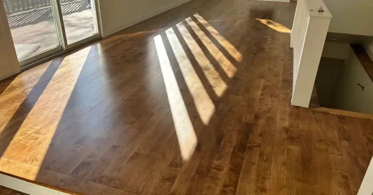 Sunlit empty hardwood room with long window shadows and a stair opening on the right