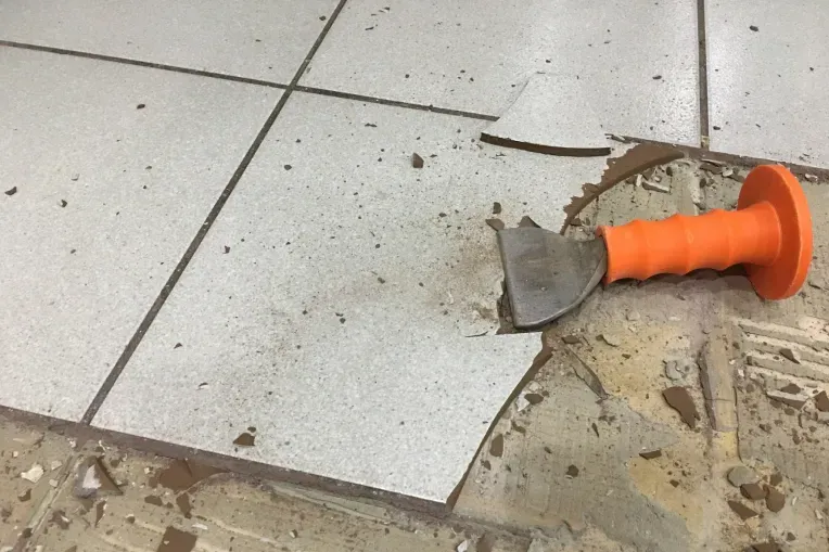 Broken floor tile with debris and an orange hand tool on a dusty tiled floor.