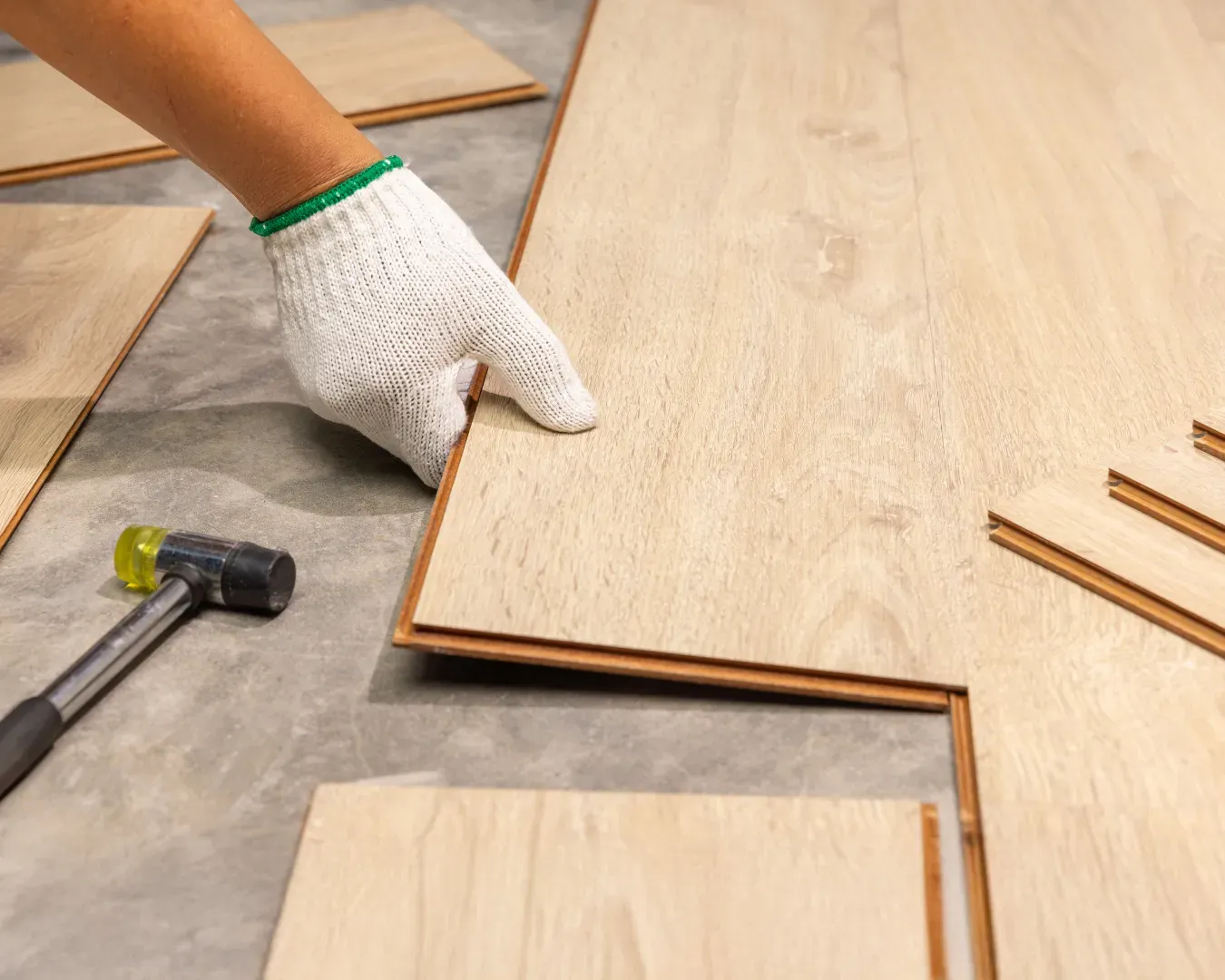 Gloved hand laying wood flooring planks with a hammer nearby