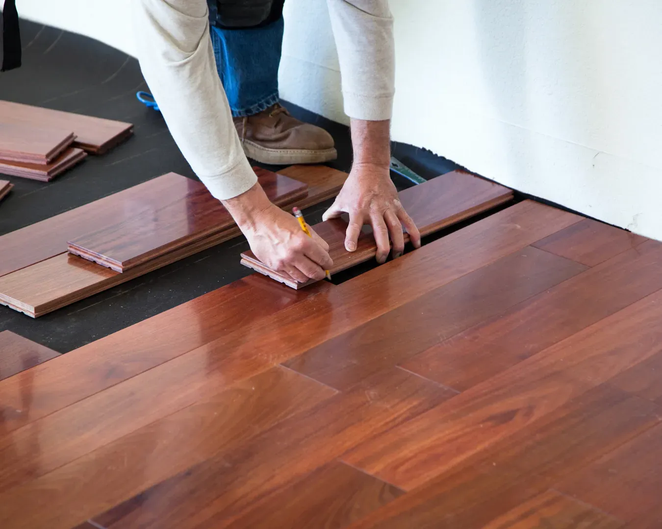 Hands installing reddish-brown hardwood flooring planks on a wooden floor