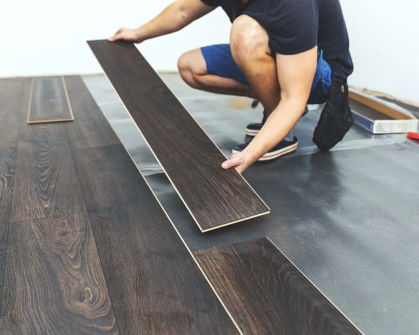 Worker installing dark wood flooring in a room with underlayment sheet visible.