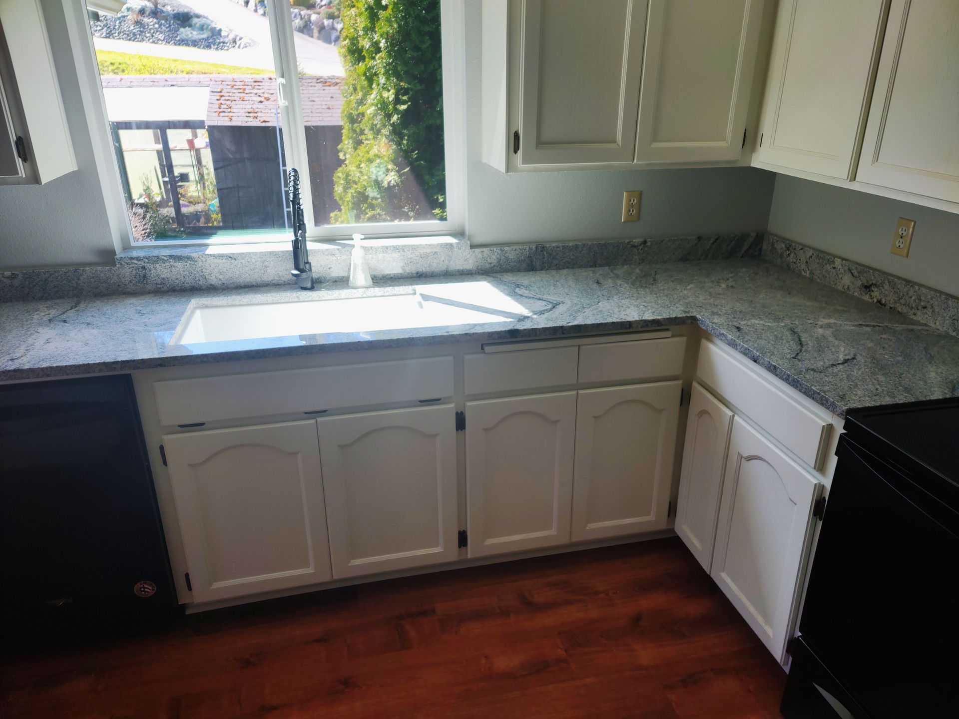 A kitchen with white cabinets , granite counter tops , a sink and a window.