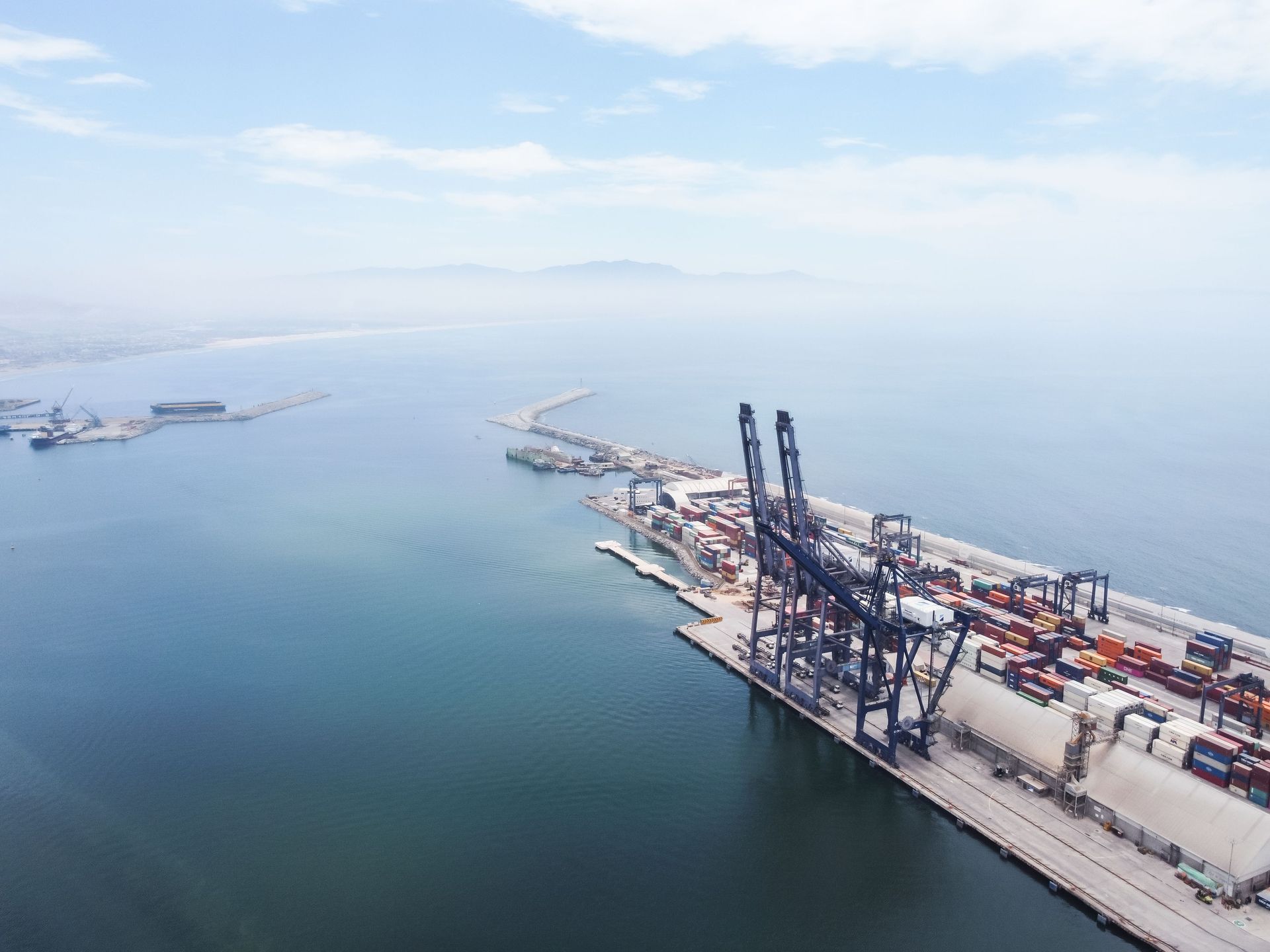 Aerial view of a cargo port with cranes, containers, and calm blue water under a hazy sky in México