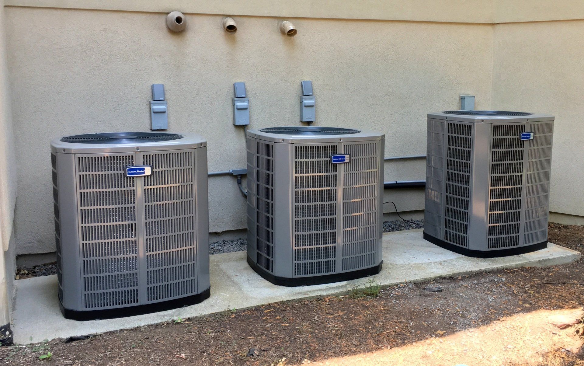 Three outdoor HVAC units beside a beige building wall on a concrete pad, with dirt and gravel in front