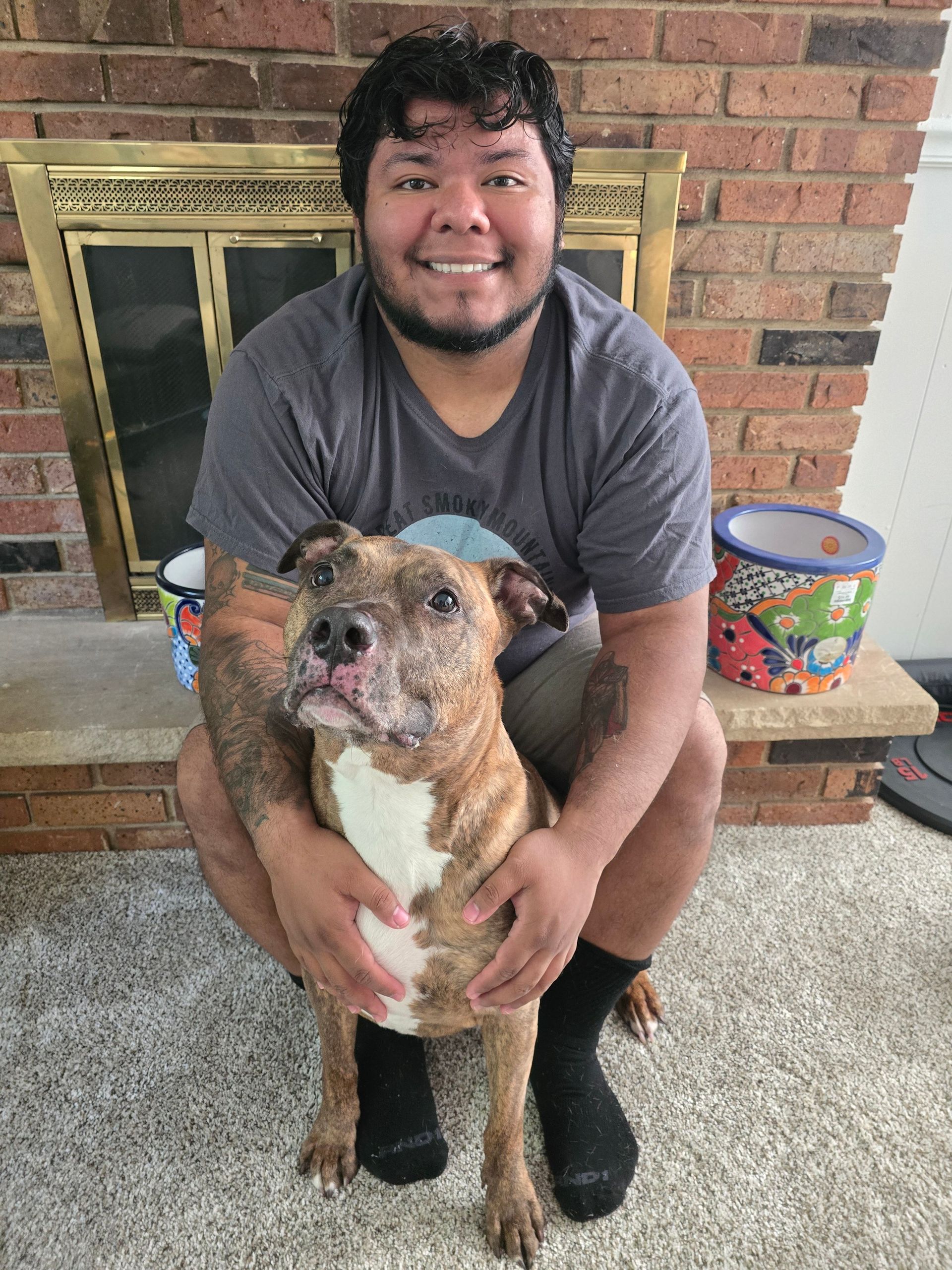 Man sitting on a brick hearth holding a brindle dog in front of a fireplace, both looking at the camera.