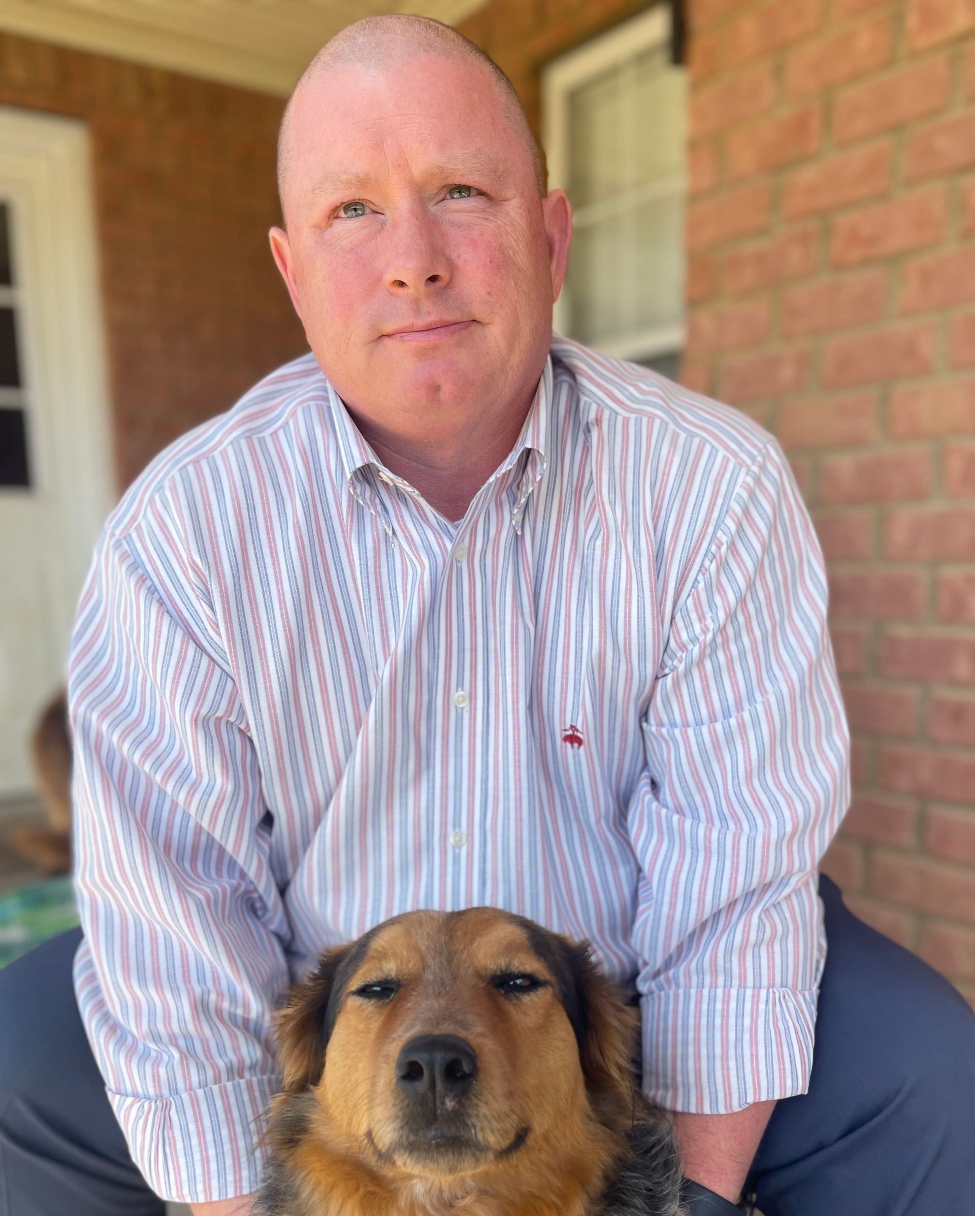 Man kneeling with a brown dog on a porch, brick wall and doorway behind them